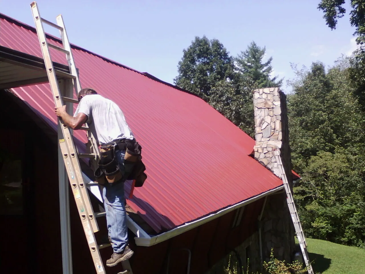 Roofer on a ladder beside a red metal roof and stone chimney — Soffit & Fascia Repair by Roofing Brothers in Baxley